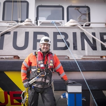 A smiling worker in safety gear and a helmet stands in front of a boat labeled TURBINE. He is wearing a harness and an orange jacket, with marine ropes and safety equipment visible around him.